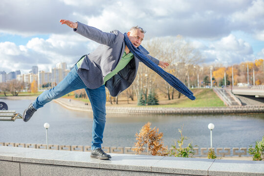 Portrait of middle-aged man balancing standing on one leg on concrete parapet near river, stretching hands in city.