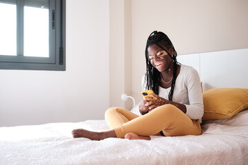 Cheerful black woman with vitiligo resting on bed while browsing cellphone