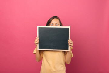 Young woman with shocked eyes is hiding behind a blackboard with copy space.