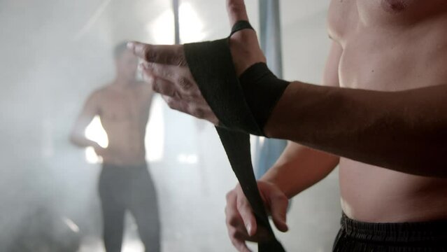 Close-up view of a boxer wrapping his hand in slow-motion with bandage in slow-motion inside the gym. Man practicing karate with nunchuck in the background