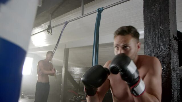 Caucasian male boxer wearing boxing gloves, training with sand bag in slow-motion at the gym while another man practicing karate with nunchuck
