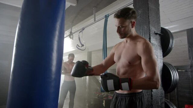 Boxer punching the boxing bag in slow-motion wearing gloves at the gym while another man practicing karate with nunchuck