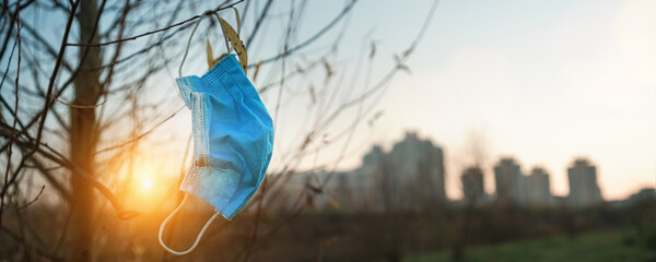 Blue old used medical face mask hanging on tree branch at autumn urban park.