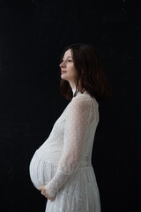 Fototapeta premium a pregnant brunette stands in profile on a black background in a white lace wedding dress hugs her big belly and looks away