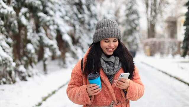 A young girl, in a pink coat, strolls through the winter park, writes a message and drinks coffee