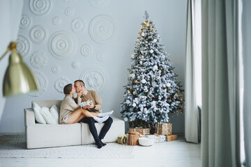 Naklejka premium A couple in love on New Year's Eve in a cozy home environment near the Christmas tree. New Year's love story. Beautiful couple in sweaters posing in studio. A guy and a girl celebrate the new year
