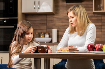 White Caucasian mom and little daughter talks while having breakfast in modern kitchen, girl holds cell phone, device. Conversation with child, upbringing,education. Parent responsibilities Motherhood