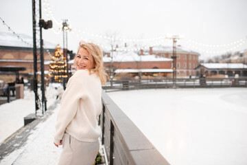 Young beautiful smiling blonde woman wearing coat standing over Christmas city decorations and glow lights at ice rink outdoor. Winter greeting holiday season.