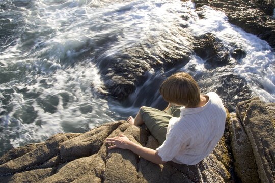 A young man sits on the rocky coast by the ocean and contemplates.