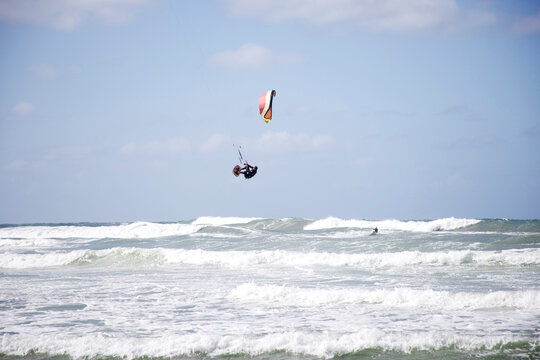 A male kitesurfer jumps and prepares to land while another male rides in the background.