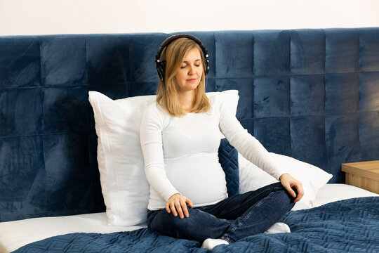 Pregnant female with headphones on head, listens to hypnosis, music,meditates,sitting on bed. Baby delivery preparation.