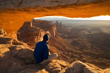 Man sitting under arch looking at desert view during sunrise, Canyonlands National Park, Utah.