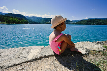 Back of baby girl is swimsuit and hat sit in pier of view beautiful Bled Lake, Slovenia.