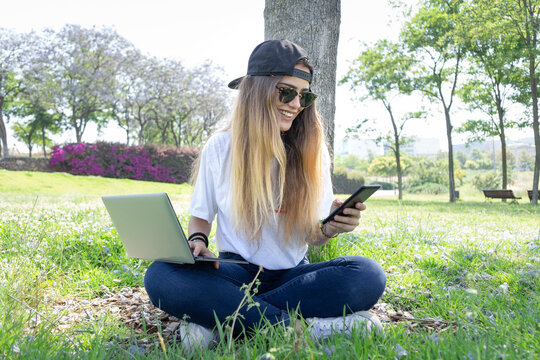 Front view of a happy student girl working with a laptop and smartphone in a green park of an university campus