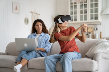Enthusiastic positive teenage girl with VR glasses for sports training with virtual baseball bat sits on couch of home. Attentive mother observed by daughter using futuristic helmet for AR video game