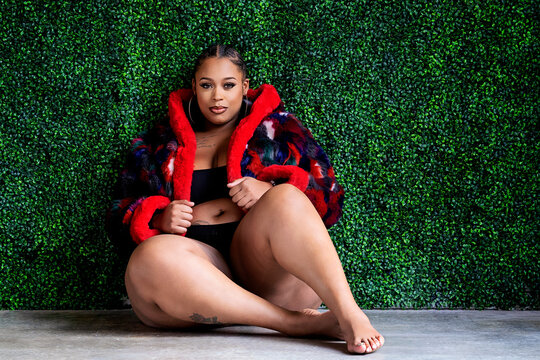 Plus size Black woman sitting in front of grass wall with coat bra and panty on