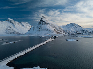 Photo depuis un drone sur une route qui traverse la mer entre les fjords en norv&egrave;ge dans un paysage enneig&eacute; au d&eacute;but de l'heure bleue au dessus du pont de Stor Buoya avec une vue sur la montagne Volan