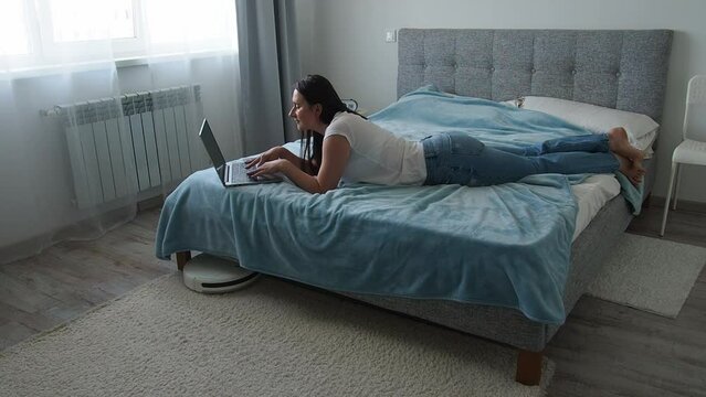 White robotic vacuum cleaner cleaning the floor while woman lying on the bed and using laptop. Smart technology concept. Woman in white t-shirt and blue jeans enjoying home cleaning. Mock up.