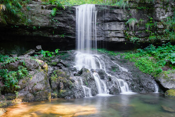 Beautiful waterfall during spring with silver colored water, cascading down the cliff covered by green plants - 550123583