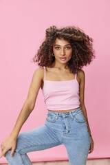Woman with curly afro hair dancing in pink top and jeans on pink background, smile, copy place