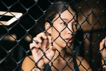 The portrait of a young Asian woman behind an aluminum mesh inside a gym. Concept of loneliness.