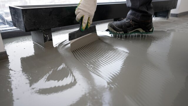 Repairman in protective gloves and uniform using metal spatula for aligning liquid self-levelling floor screed. Worker spreading liquid cement solution on construction site. 