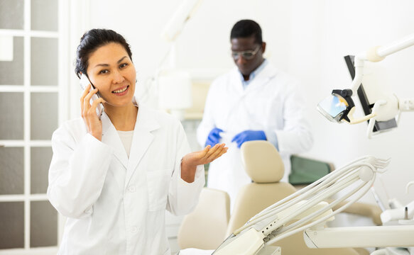 Smiling asian female dentist in white coat talking on mobile phone in modern dental office..