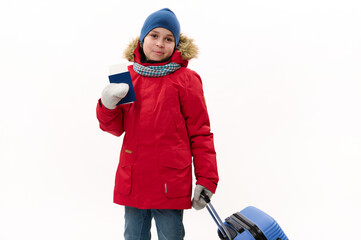Isolated portrait on white background of multi-ethnic hcild boy in warm blue parka and wool mittens, going for winter holidays, walking with air ticket and suitcase. Rest getaway. Travel concept
