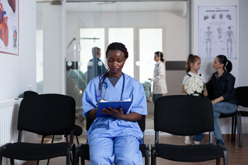 African american nurse filling out patient paperwork sitting at medical tower reception chairs. Young doctor writing prescriptions in hospital waiting room. Medical staff member reviewing patient list