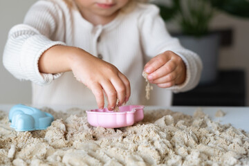 Unrecognizable cute happy caucasian,blonde,curly-haired toddler,baby girl playing with kinetic sand indoors.Child hands close-up.Preschool kid early development,motor skill, sensorics concept.