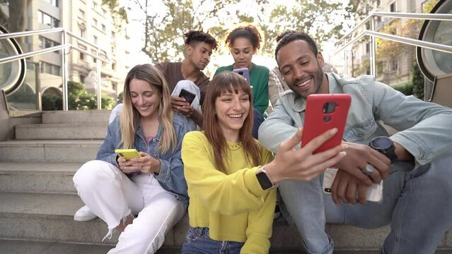 Happy mixed race group of University colleagues sitting on stairs outdoors in the city, using mobile to watch funny videos online, laughing and having fun together. Generation addicted to technology.