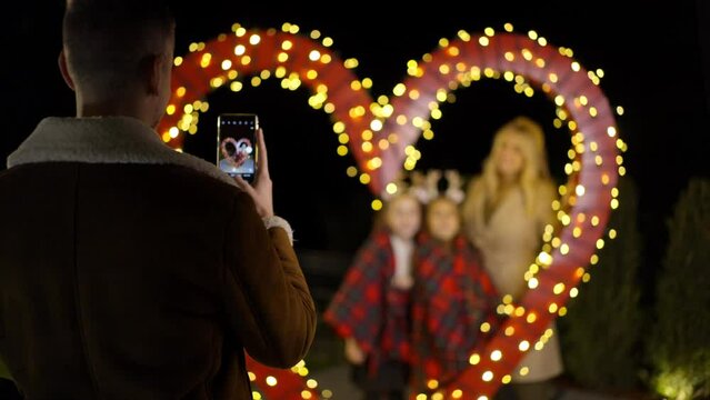 Young man is taking a picture of his family standing behind the heart shaped Christmas decoration. Slow motion. 