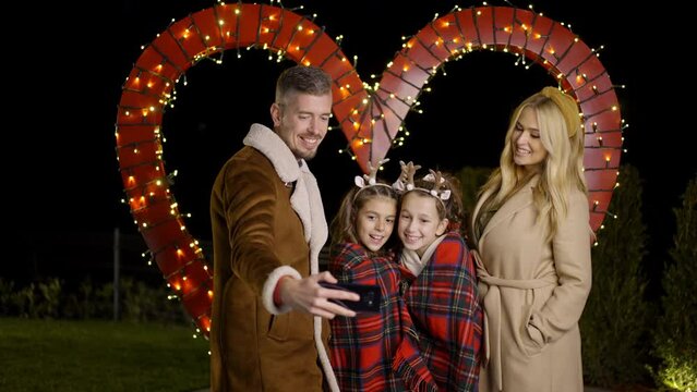 Young family taking a selfie, while standing behing heart shaped Christmas decoration. Slow motion. 