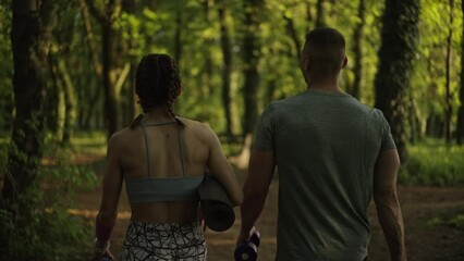 Back view of two people talking and walking in the nature after training, holding yoga mat and dumbbells.Healthy lifestyle concept. Slow motion. 