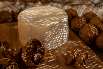 Composition of a round head of cheese with white mold and walnuts on a bamboo board on a background of burlap, with droplets of light penetrating through the fabric