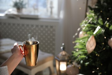 Woman with golden cup of hot drink near Christmas tree at home, closeup