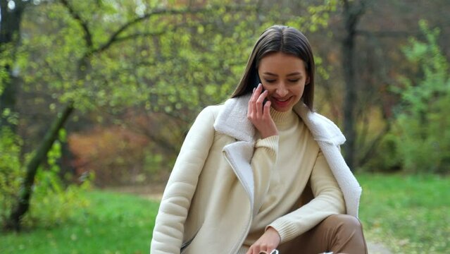 Elegant brunette in white jacket sitting outdoors in autumn. Lady speaks on the phone smiling broadly. Close up. Blurred backdrop.