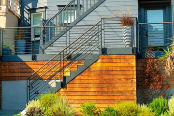 Modern stairs with black metal hand railing with horizontal natural wooden slats with plants and foliage foreground and stucco gray wall background - Powered by Adobe