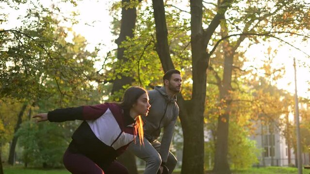 Young beautiful woman and handsome man swinging dumbbells in the sunny park. 
