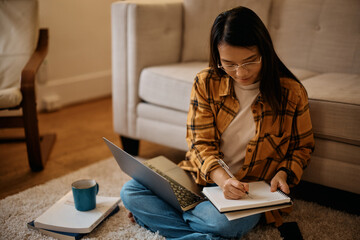 Obraz premium Young Asian woman using laptop and taking notes while studying at home.