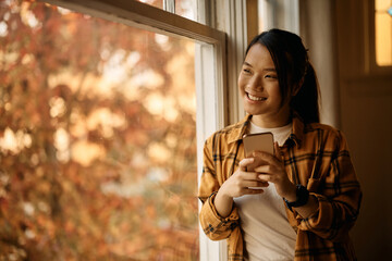 Happy Asian woman using mobile phone by window at home.