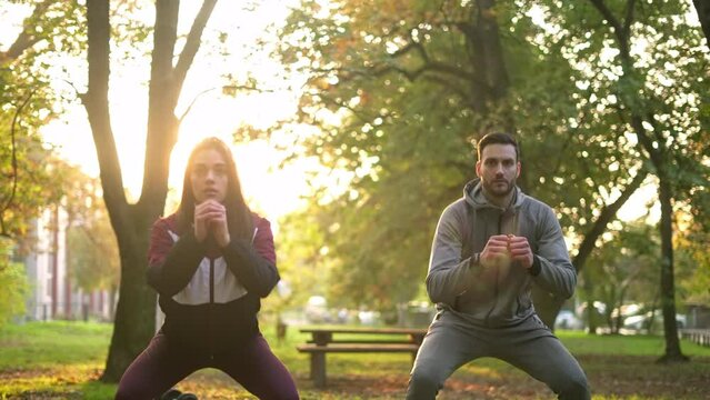 Young beautiful woman and handsome man doing squatting exercise in the sunny park. 