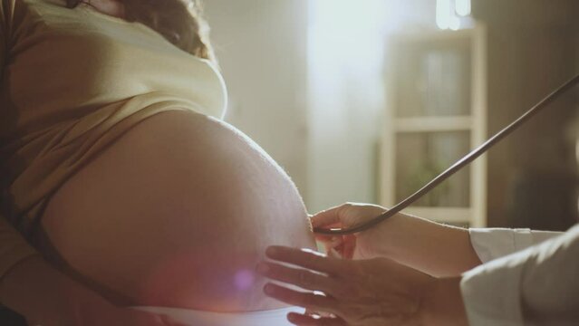 Close-up view of a female doctor using stethoscope to examining young pregnant woman's belly in clinic. Gynecology concept