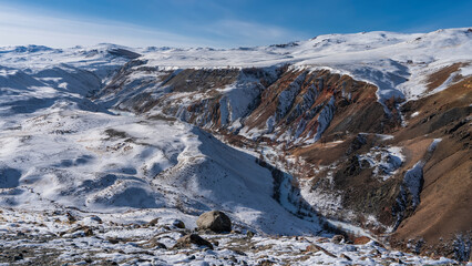 The river winds through a gorge between the red-brown slopes of the mountains. Scattered rocks are...