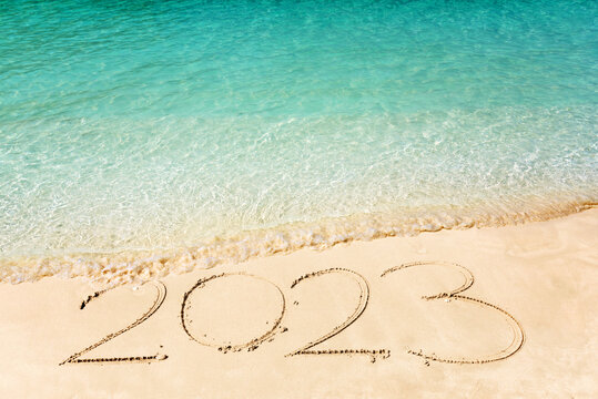 Happy New Year 2023, lettering on the beach with wave and clear blue sea. Numbers 2023 year on the sea shore, message handwritten in the golden sand on beautiful beach background. New Year concept.