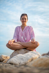 Naklejka na ściany i meble Happy relaxed woman meditation in lotus position practicing yoga sitting on stone beach sea waves