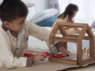 Cute boy playing with wooden educational toy house, using toy hammer to fix while sister playing on the bed in the blurry background.