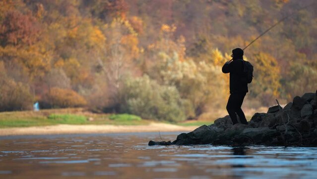 Silhouette of man fishing and throwing a spinning rod into the lake in autumn. View from the lake.