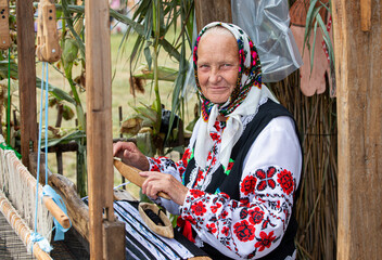 An old Belarusian or Ukrainian woman in an embroidered shirt at a vintage loom. Slavic elderly woman in national ethnic clothes.