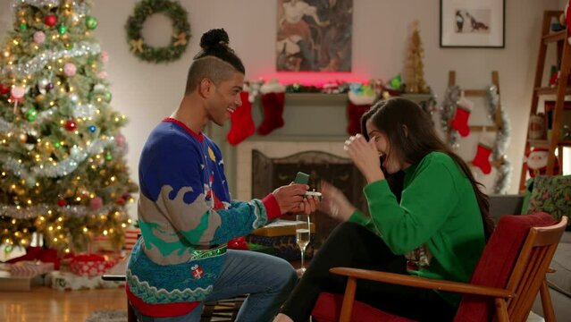 Man wearing Christmas sweater proposes to his girl friend in their living room